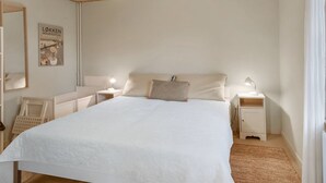 Un dormitorio con techo de madera, una cama grande con cubrecama blanco, dos mesitas de noche con lámparas, un tocador y un espejo montado en la pared.