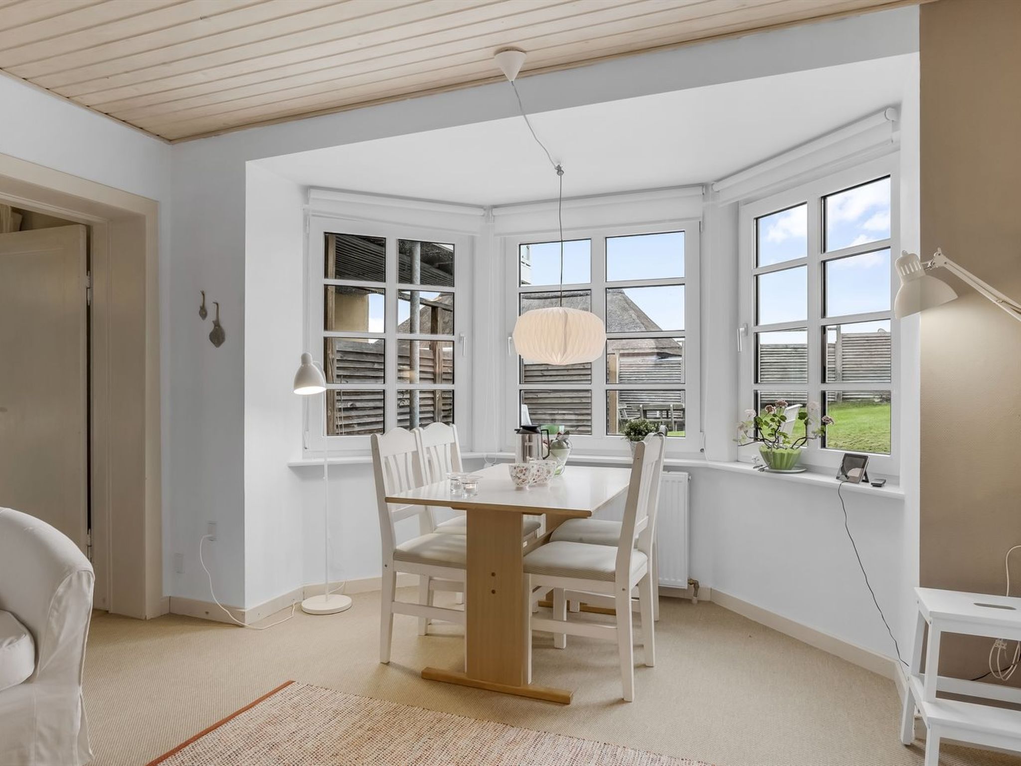 Et spiseområde med et træbord og stole, store vinduer med skodder samt en hængende lampe.