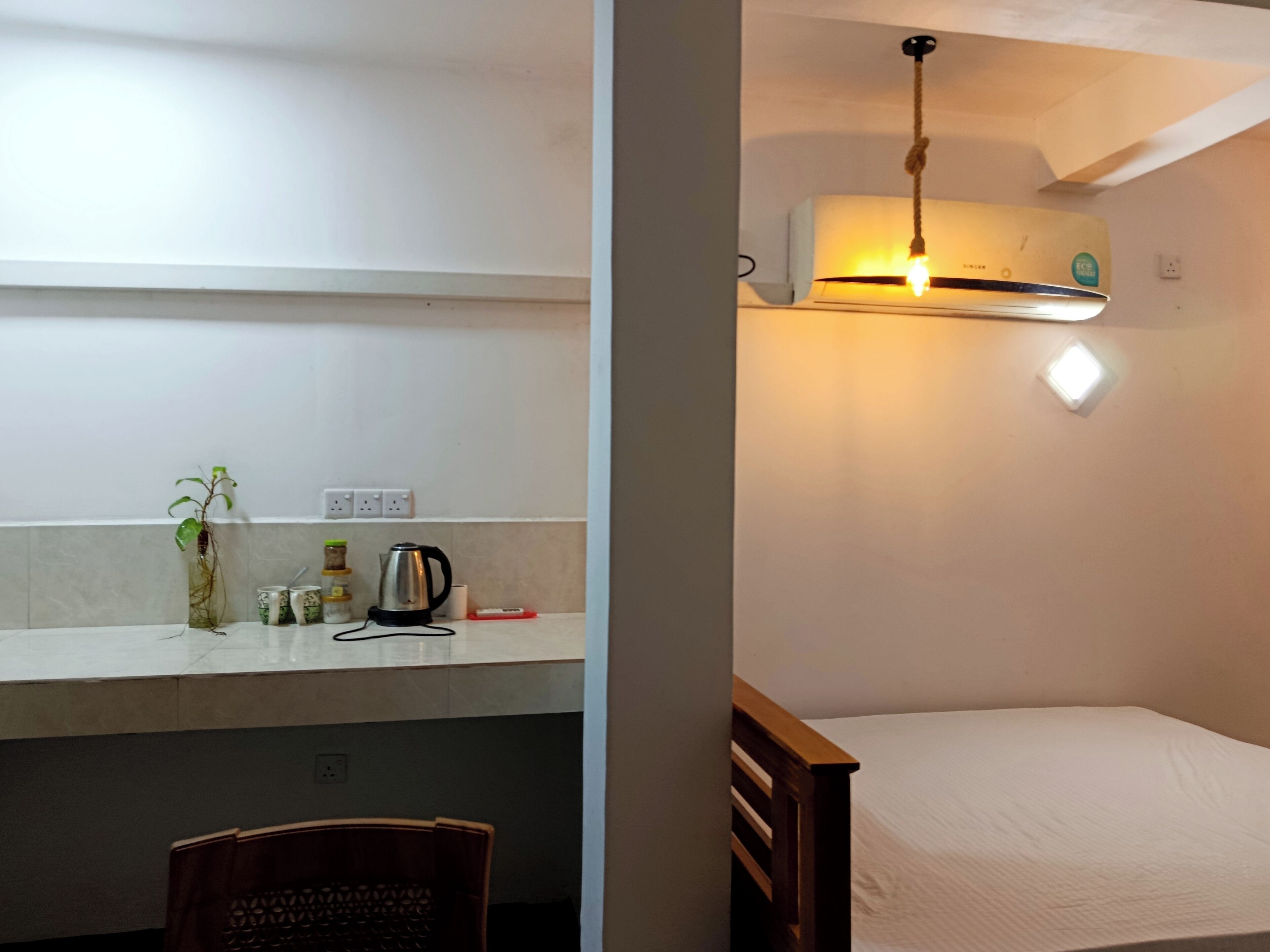 A small, simple room with a bed, a desk with a chair, and a kitchenette area with a kettle and jars.