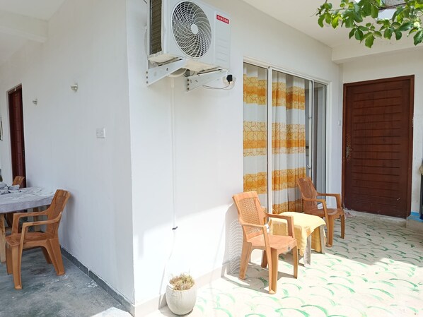 Egy szoba, melynek van egy fehér fala, egy mennyezeti ventilátora, egy tolóüveg ajtaja és egy barna ajtaja. Két fa szék és egy kis, virágos cserép áll benne.