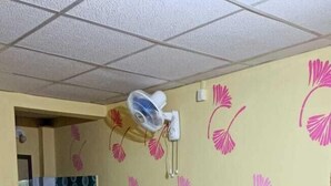 Quarto com cama, ventoinha na parede, pia pequena e lixeira.