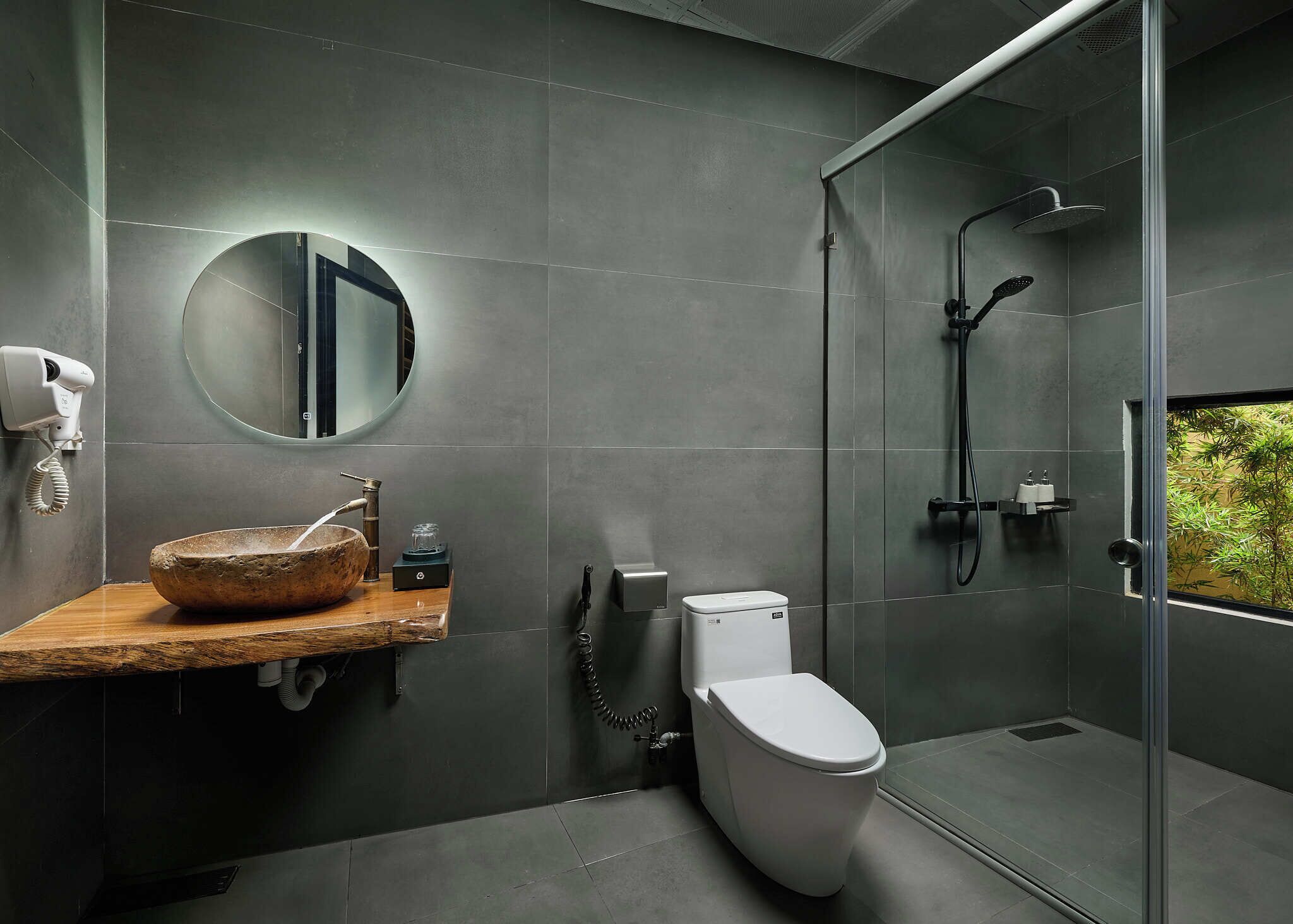 A modern bathroom with a round mirror, a wooden sink, a toilet, and a glass-enclosed shower area.