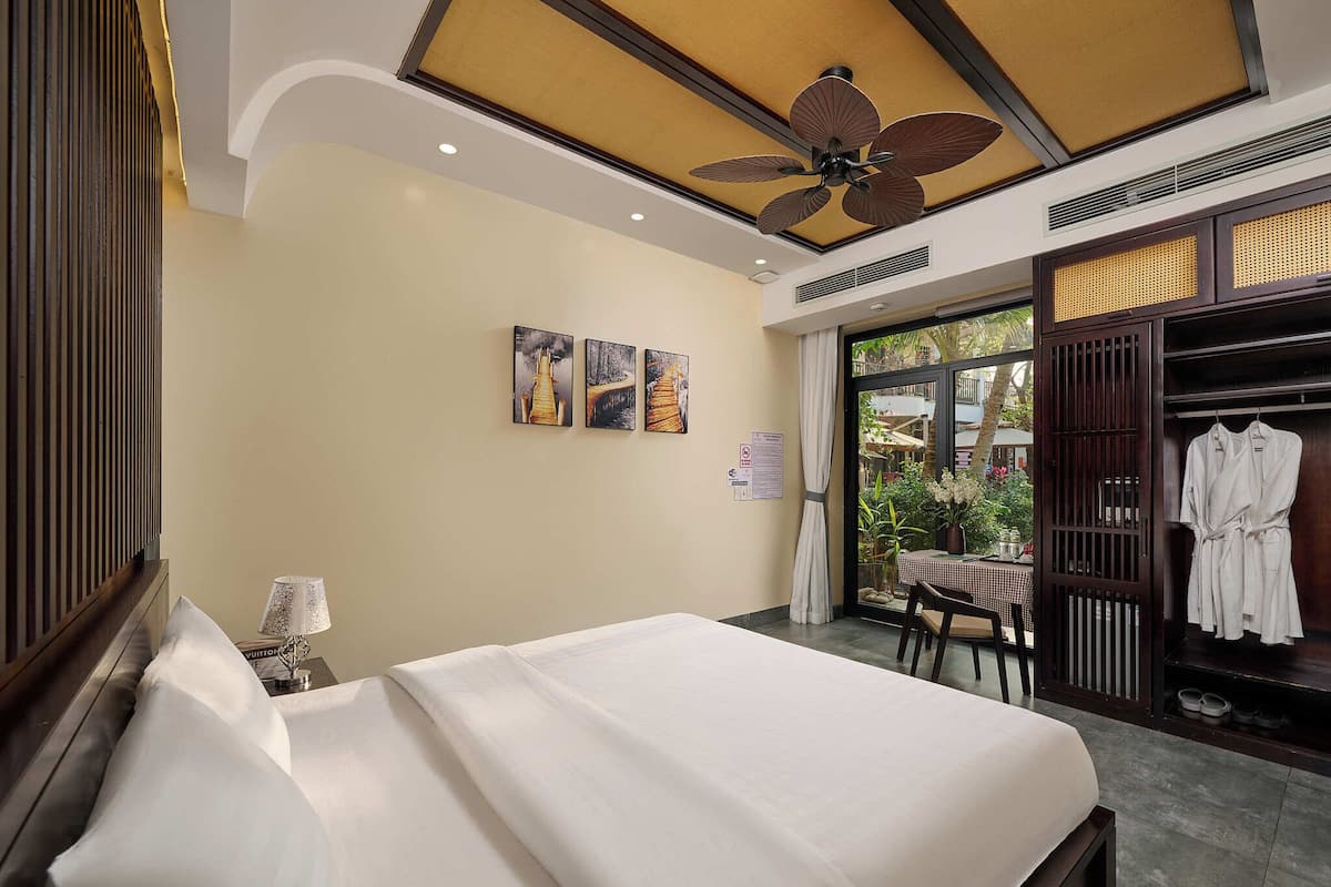 A hotel room with a bed, a ceiling fan, artwork on the wall, a table with chairs, and a view of the outside.
