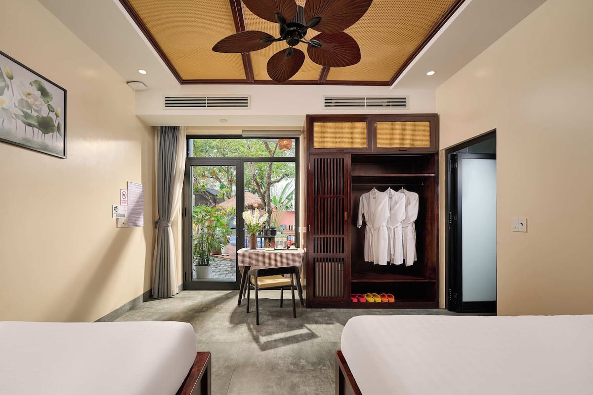 A hotel room with two beds, a dining table, a wardrobe, and a ceiling fan.