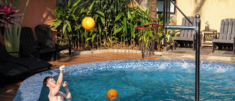 A swimming pool with a person playing with a ball, surrounded by plants and chairs.