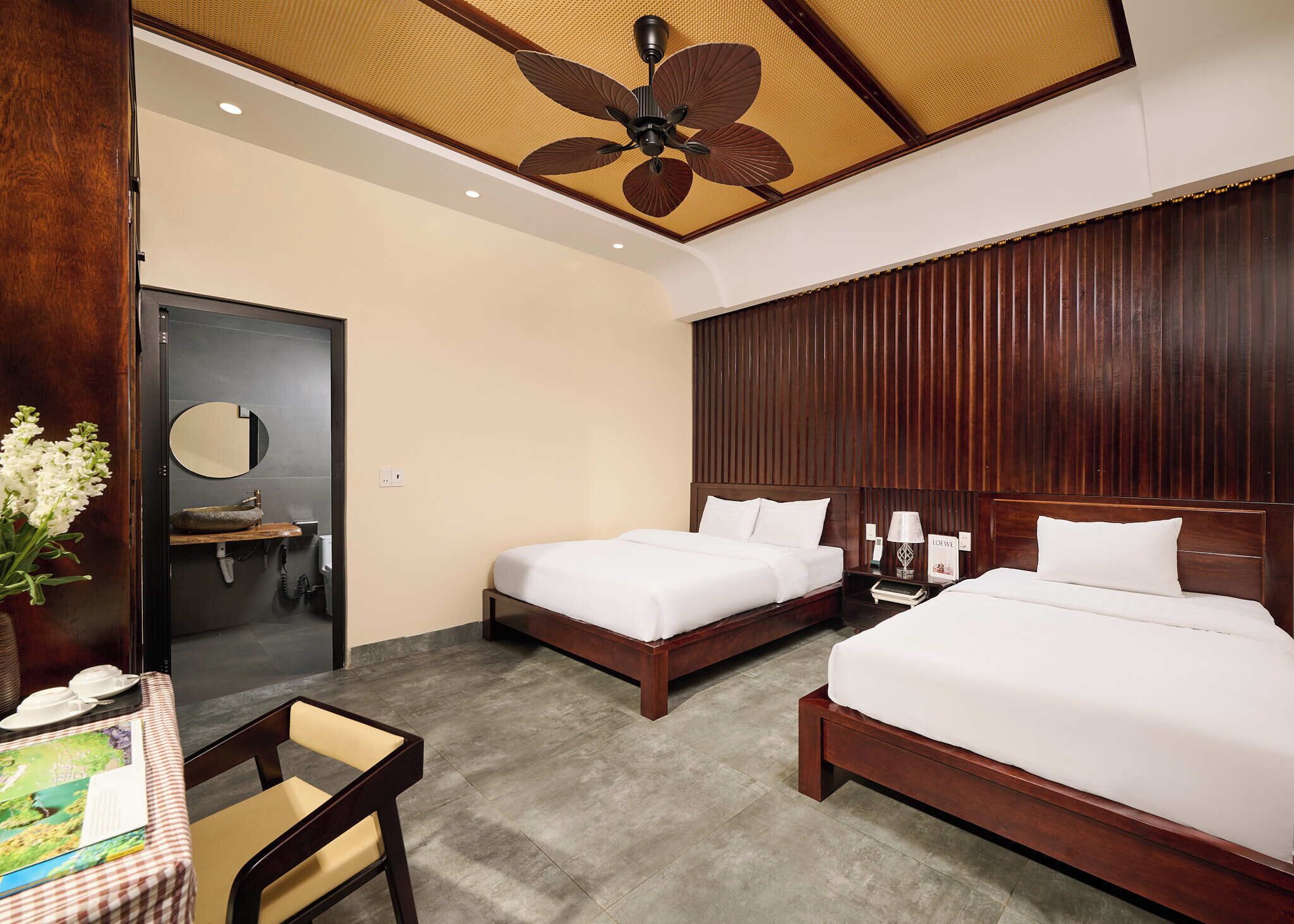 A hotel room with two beds, a wooden headboard, a ceiling fan, a round mirror, a small sink, and a table with a chair.