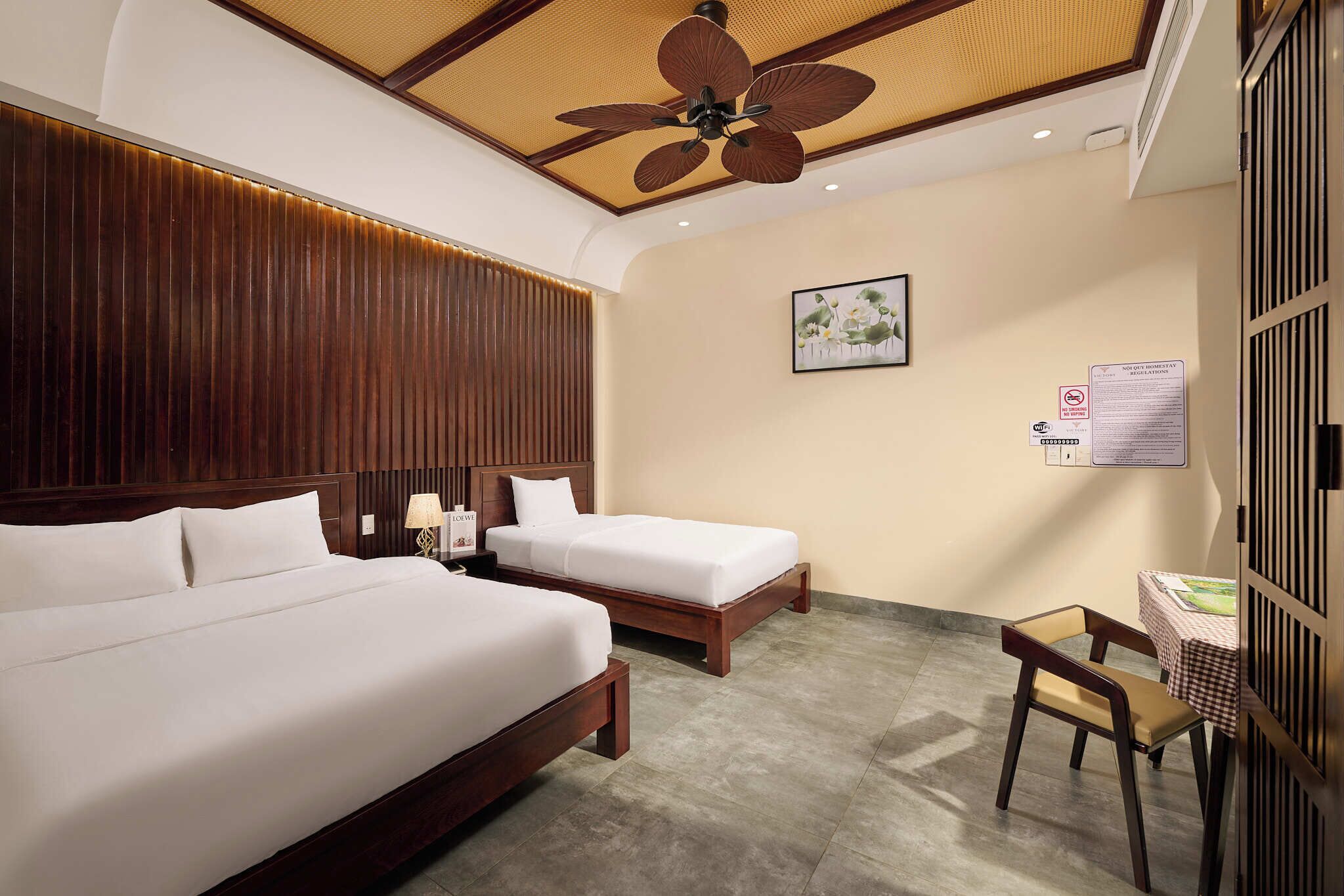 A hotel room with two beds, a ceiling fan, a wooden headboard, a small table with a chair, and a wall-mounted sign.