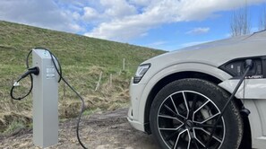 Ein weißes Auto parkt neben einer Ladestation für Elektrofahrzeuge auf einem Feldweg, im Hintergrund ist ein grasbewachsener Hügel zu sehen.