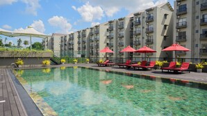 Une piscine aux eaux cristallines, bordée de chaises longues rouges et d’ombrelles, le tout entouré d’un immeuble à plusieurs étages doté de balcons.