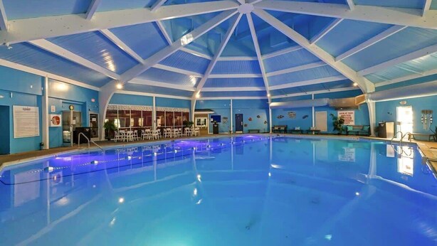 An indoor swimming pool with a blue ceiling and walls, surrounded by seating areas and a bar. - Game Room, Hot Tub, Fire Pit, Dogs OK, Indoor Pool (Long Pond)