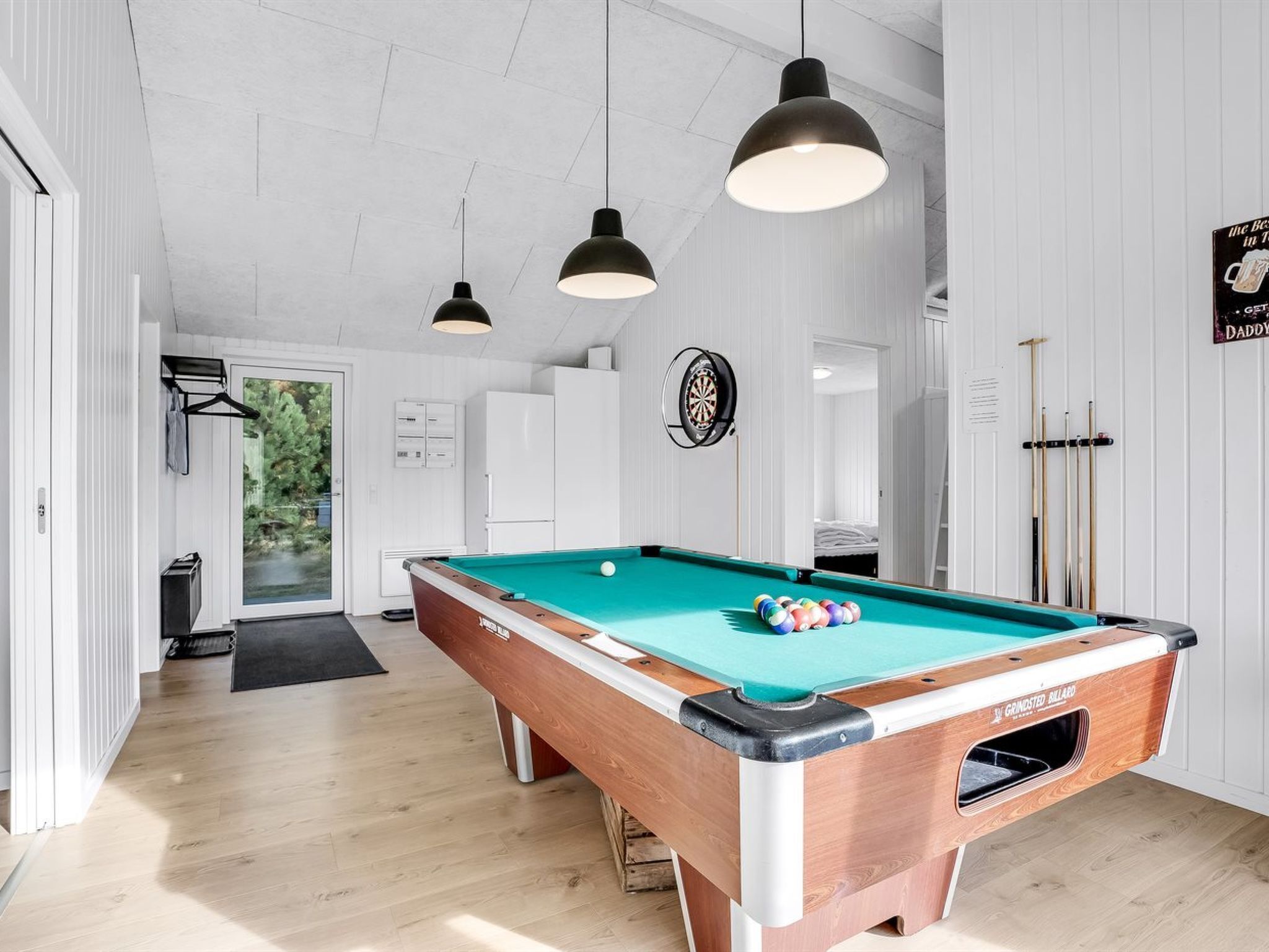A pool table in a room with white walls, a dartboard, and pool cues.