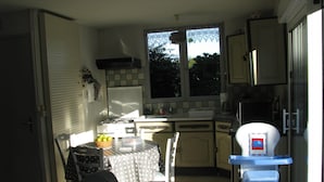 A compact kitchen with a dining area, a window with lace curtains, and a small table with a polka dot tablecloth. - Gîte 10 minutes from the beach (Hudimesnil)
