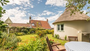 A garden with a thatched-roof house, a wooden bench, and a table with a polka dot tablecloth.