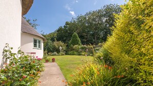 A white house with a thatched roof, a paved pathway, and a well-maintained garden with various plants and flowers.