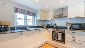 A modern kitchen with white cabinets, a stainless steel range hood, a built-in oven, a microwave, and a window with striped valance.