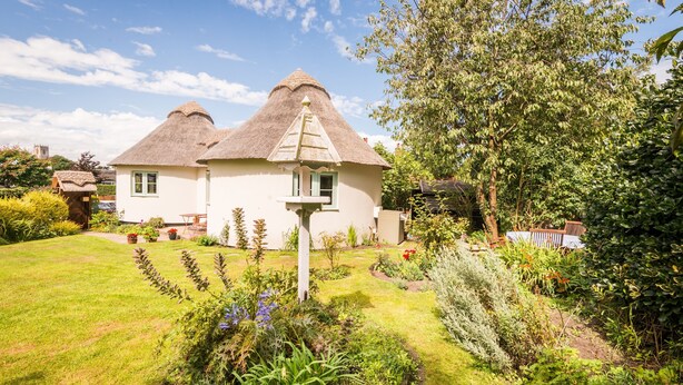 A house with two thatched-roof structures, surrounded by a well-maintained garden with various plants and a small fountain.