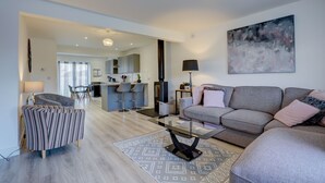 Un salon moderne avec un canapé gris, un fauteuil à rayures et une table basse en verre. On aperçoit une cuisine à l’arrière-plan, avec des tabourets de bar.