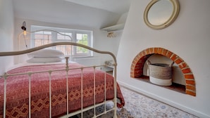 A bedroom with a white bed, a red patterned bedspread, a white wall with a round mirror, and a brick fireplace. - Rose Cottage | Dog-Friendly Village Stay, 2 Minutes from the Beach (Winterton-on-Sea)
