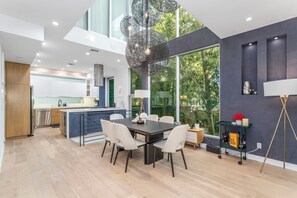 A modern kitchen with a central island, a dining area with a dark table and white chairs, and a large window providing a view of greenery.