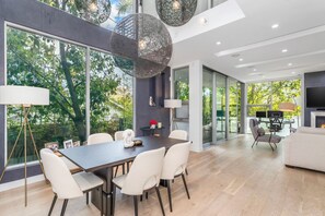 A modern dining area with a dark wooden table, white chairs, and large windows offering a view of greenery. - Beautiful home in Prime Weho w/Private Rooftop Spa! Incl. Parking/Laundry/Wifi (Los Angeles)