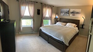 A bedroom with a bed, two windows with curtains, a bedside table with a lamp, and two wall-mounted paintings.
