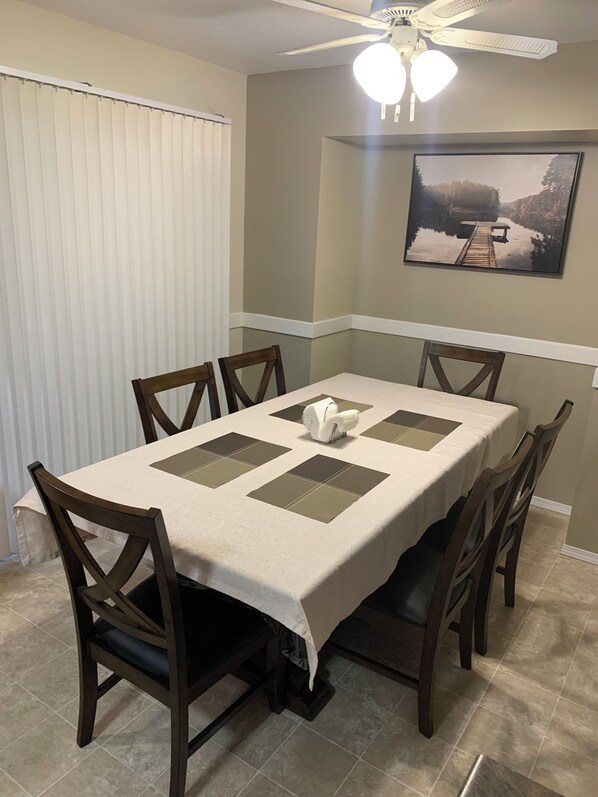 Un comedor con una mesa dispuesta para seis personas, rodeada de sillas de madera. Un ventilador de techo se encuentra arriba y un cuadro enmarca la pared.
