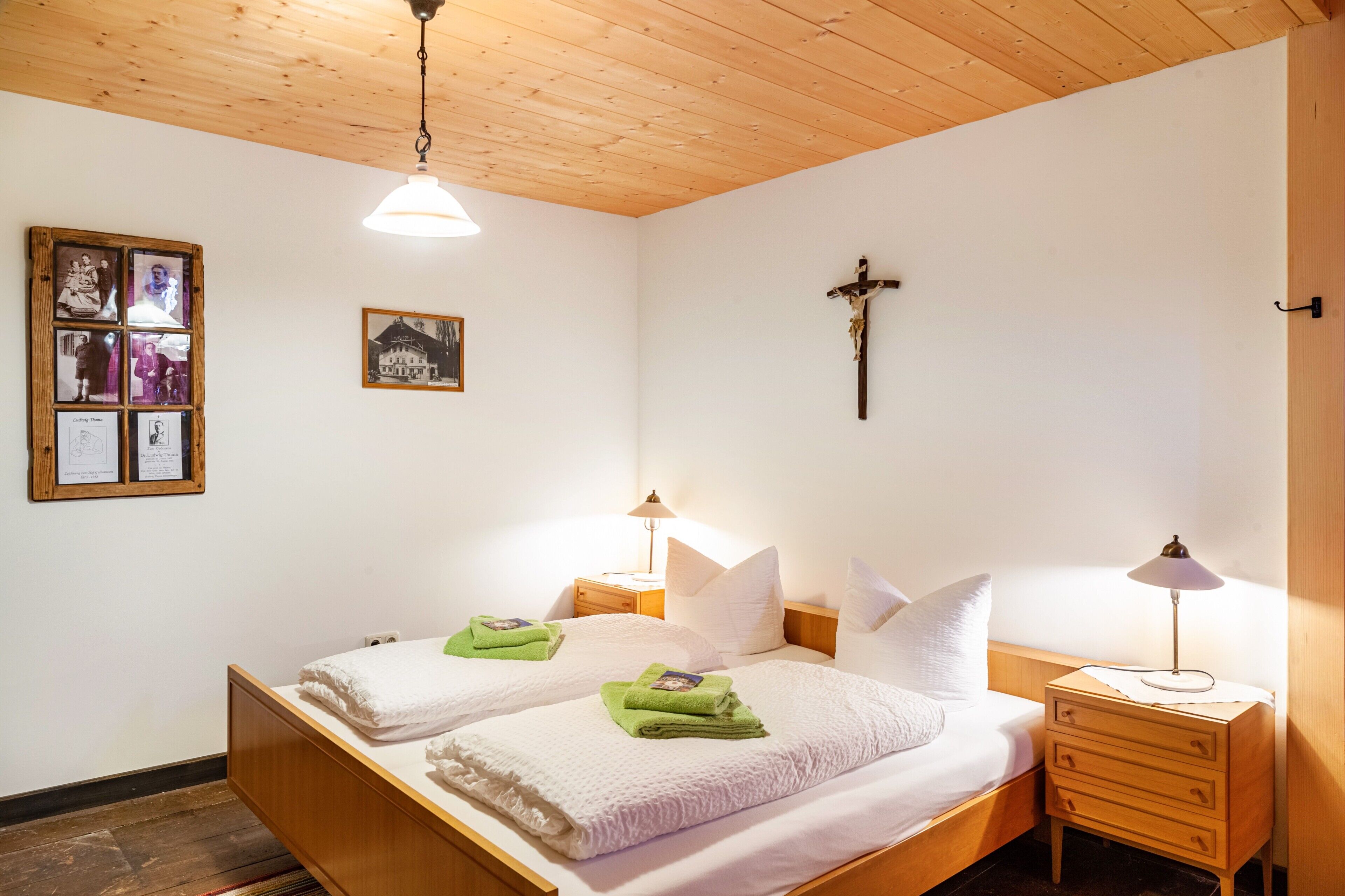 A double bed with white linens, two bedside tables with lamps, a wooden ceiling, and a cross on the wall.