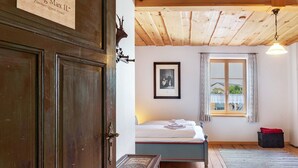 A wooden ceiling, a bed with a white mattress, a framed picture on the wall, a window with curtains, and a small black box on the floor. - Guest Rooms "König-Max-ll" and "Ludwig-Thoma" (Oberammergau)