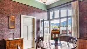 A room with a round wooden table, chairs, a wooden cabinet, a door, and a window with a view of mountains. - Guest Rooms "König-Max-ll" and "Ludwig-Thoma" (Oberammergau)