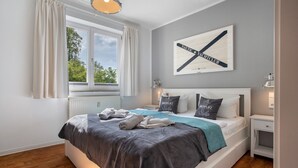 A neatly made bed with a grey and blue bedspread, a window with curtains, a wall-mounted artwork, and a bedside table with a lamp. - Holiday Apartment 'Inselliebe Rügen' with Balcony, Ahoi Pool & Sauna Included (Sellin)
