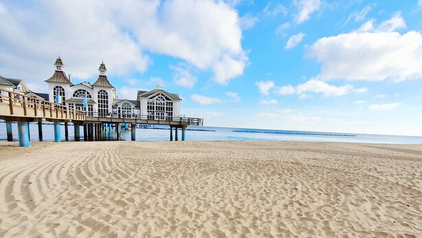 A beach with a pier and a building with multiple windows and a flag. - Holiday Apartment 'Inselliebe Rügen' with Balcony, Ahoi Pool & Sauna Included (Sellin)