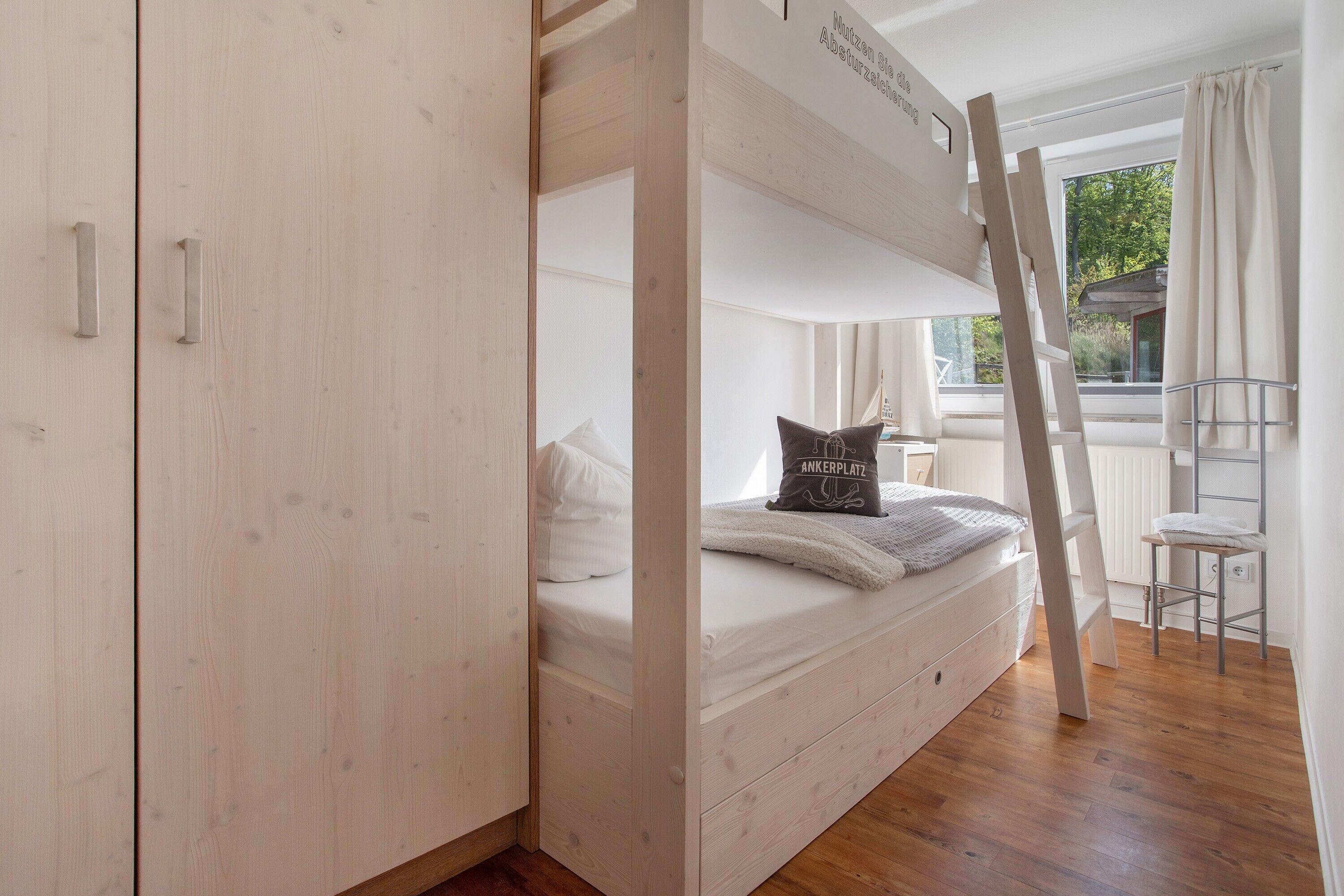 A bunk bed with a ladder, a wooden wardrobe, and a window with a view of greenery.