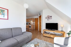 Un salon moderne avec un canapé gris, un téléviseur à écran plat posé sur un meuble en bois, et une cuisine en arrière-plan.