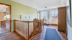 Ein Zimmer mit Holzbalkon, einem Esstisch, einem Fenster mit Vorhängen und Blick auf Bäume.
