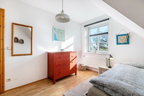 Une chambre avec une commode en bois, un lit avec une literie à motifs, une fenêtre donnant sur un paysage agréable, une carte accrochée au mur et un cœur décoratif suspendu.
