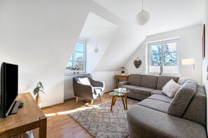 Un appartement moderne de style « loft », avec un plafond en pente, une grande fenêtre, un canapé, une table basse et un téléviseur posé sur une table en bois.
