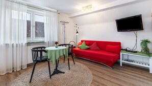 Una sala de estar con un sofá rojo, un televisor montado en la pared, una mesita con un jarrón y una ventana con cortinas.