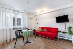 A living room with a red sofa, a TV mounted on the wall, a small table with a vase, and a window with curtains.