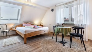 Un dormitorio con cama de madera, una mesita con mantel verde, una silla, un reloj en la pared y una ventana con cortinas.