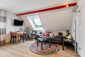 Un espace de vie de style « loft », avec un plafond en pente, un téléviseur fixé au mur, un coin repas comprenant une table et des chaises, un canapé avec des coussins et une petite table d’appoint.