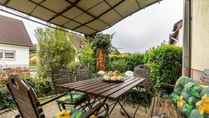 Un porche techado con muebles de madera, incluyendo una mesa y sillas, rodeado de follaje y un árbol ornamental.