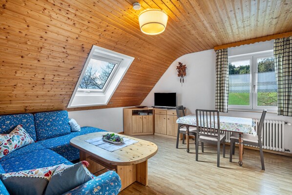 A cozy room with a wooden ceiling, a sofa, a dining table with chairs, a TV, and a window with curtains.