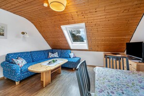 A cozy room with a wooden ceiling, a blue sofa, a wooden table, a TV, and a window.