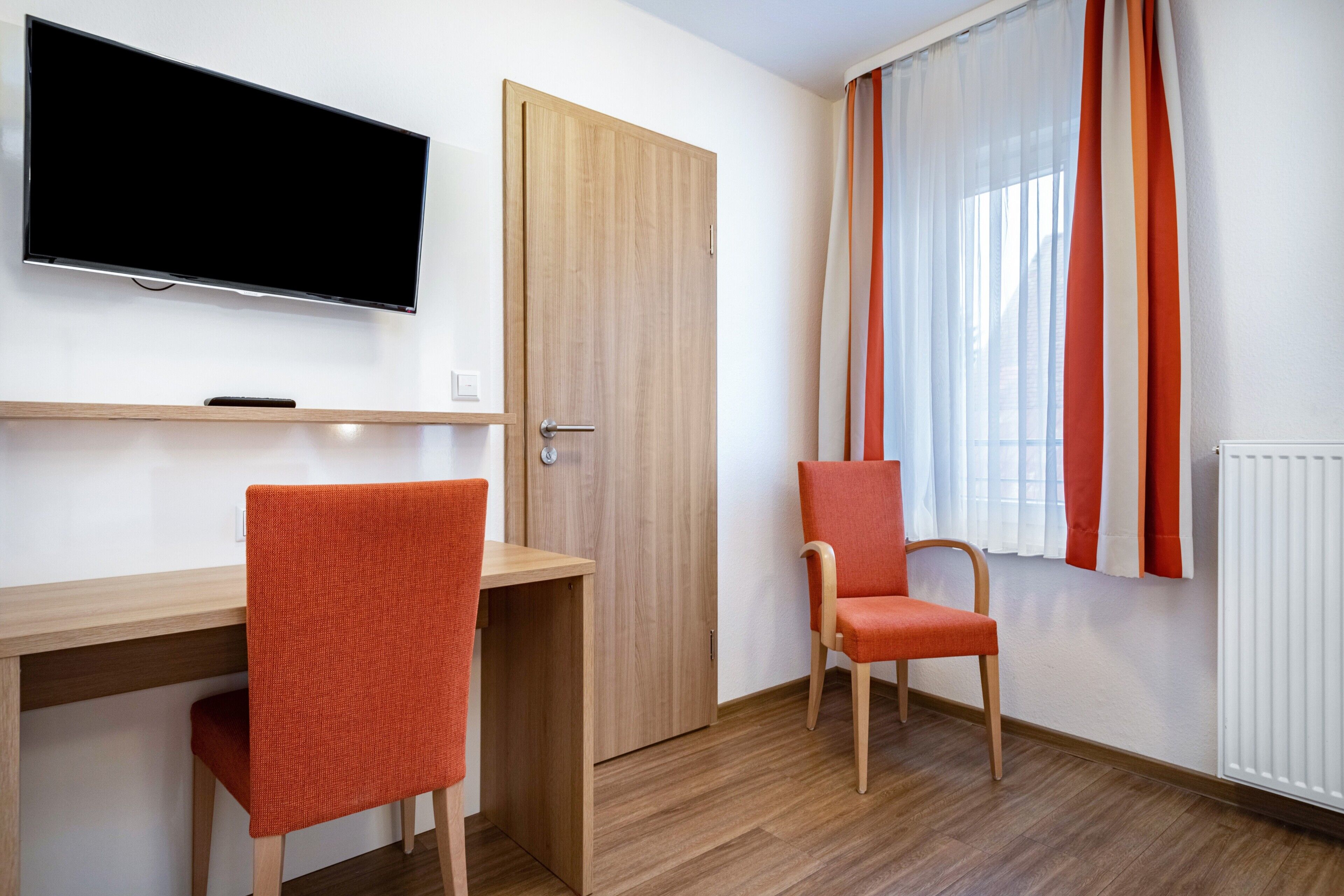 Ein Zimmer mit einem Holztisch, zwei orangefarbenen Stühlen, einem Fernseher, einer Tür und einem Fenster mit Vorhängen.