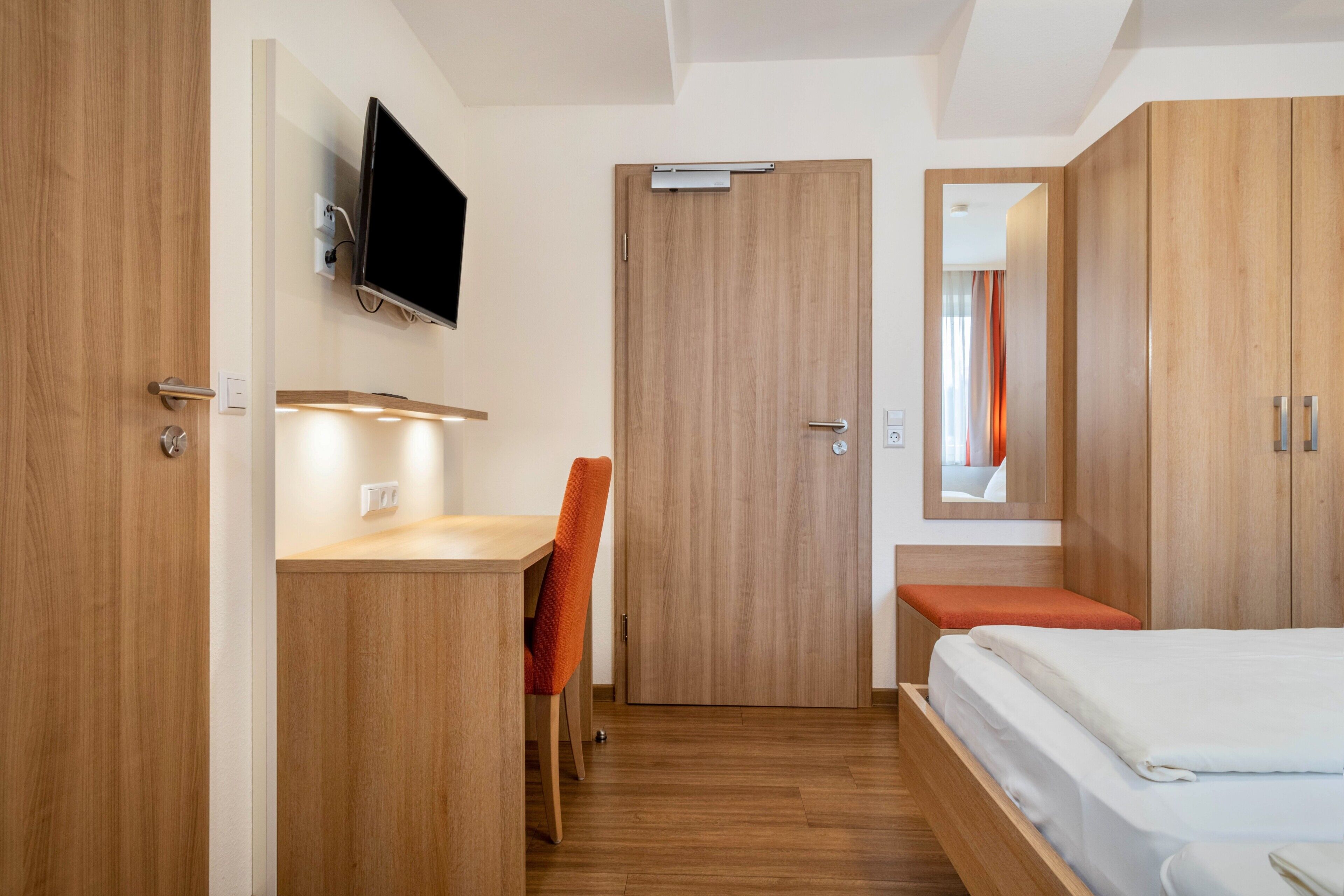 A hotel room with a wooden desk, a chair, a bed, a TV mounted on the wall, and a mirror above the bed.