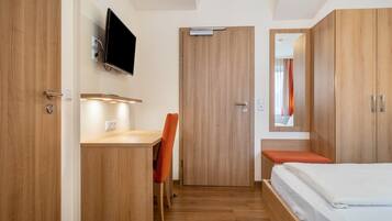 A hotel room with a wooden desk, a chair, a bed, a TV mounted on the wall, and a mirror above the bed.