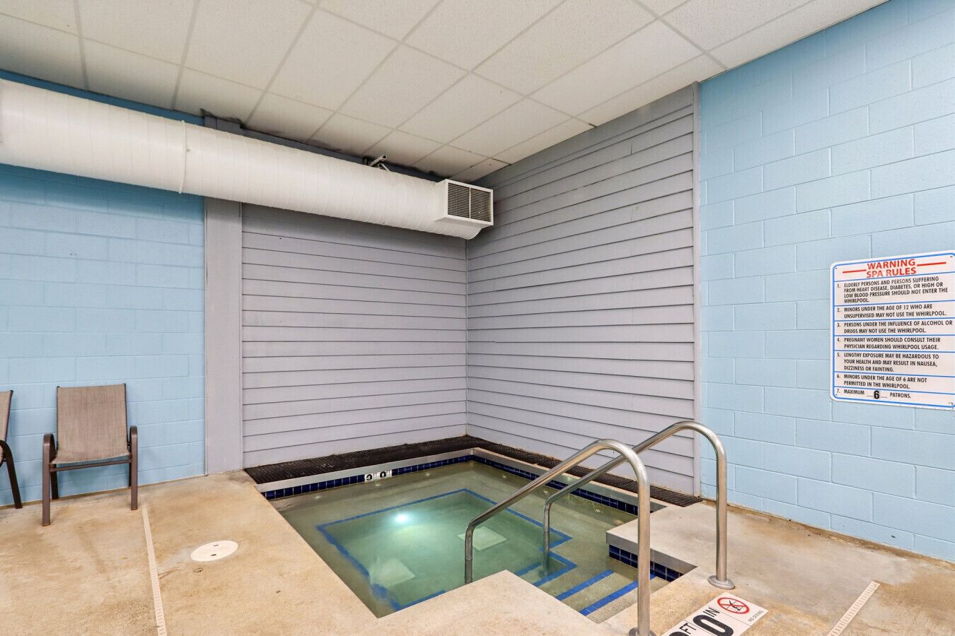 A small indoor pool with a blue tiled wall, a metal ladder, and a warning sign about spa rules.