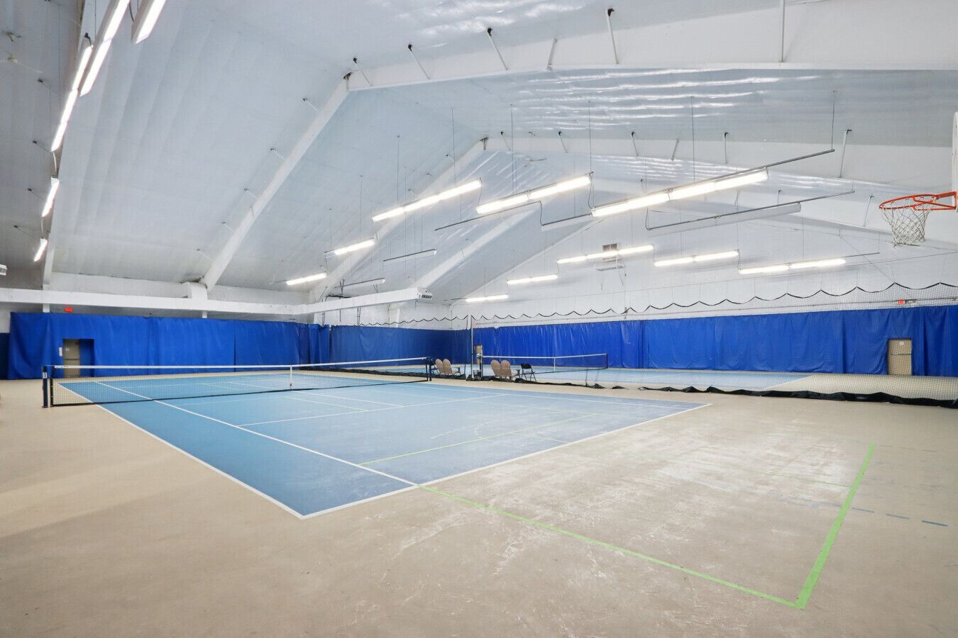 An indoor tennis court with a blue playing surface, white net, and a basketball hoop.
