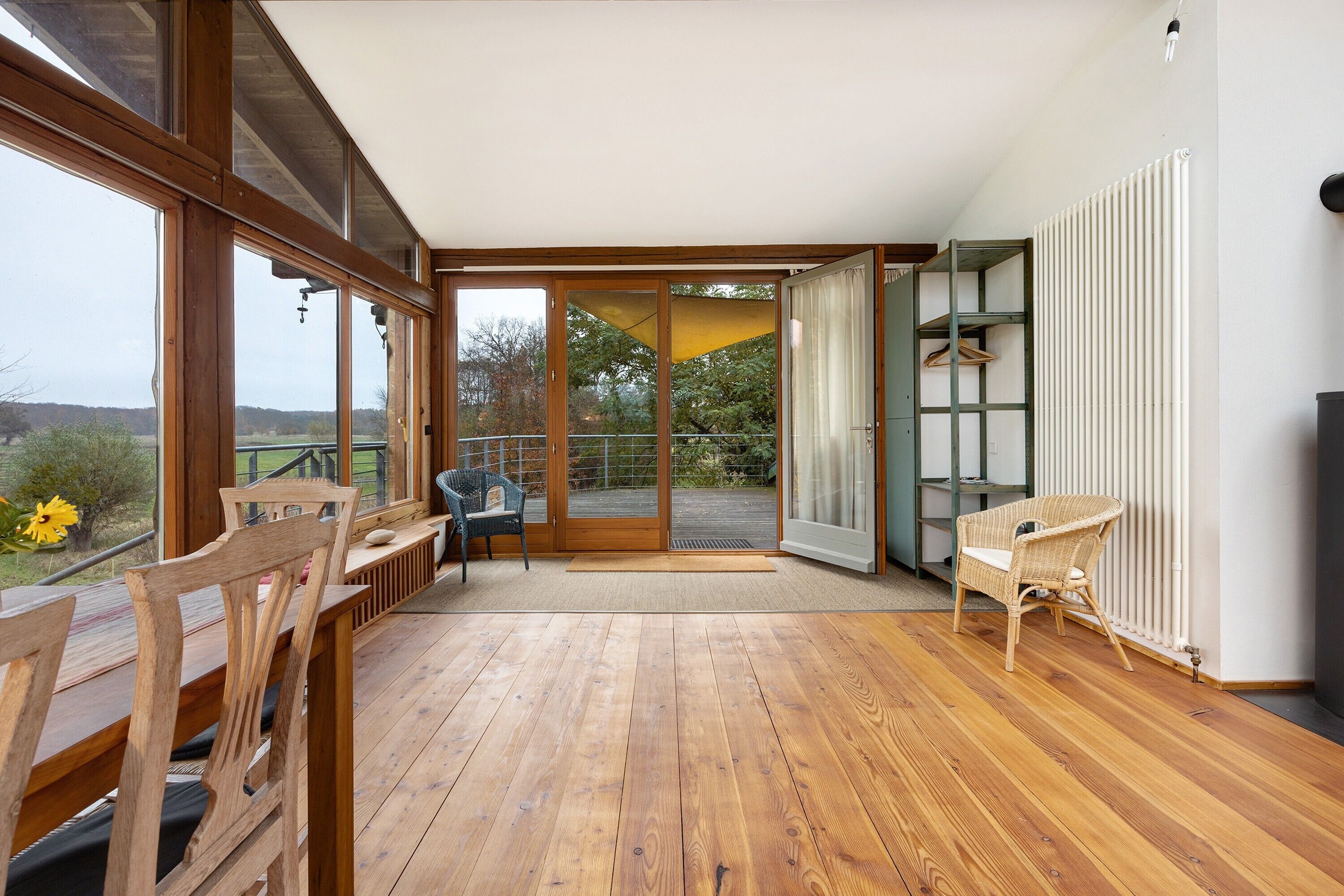 Una habitación amplia con piso de madera, ventanas grandes, una puerta corredera de vidrio que da a un espacio exterior y una silla de mimbre.