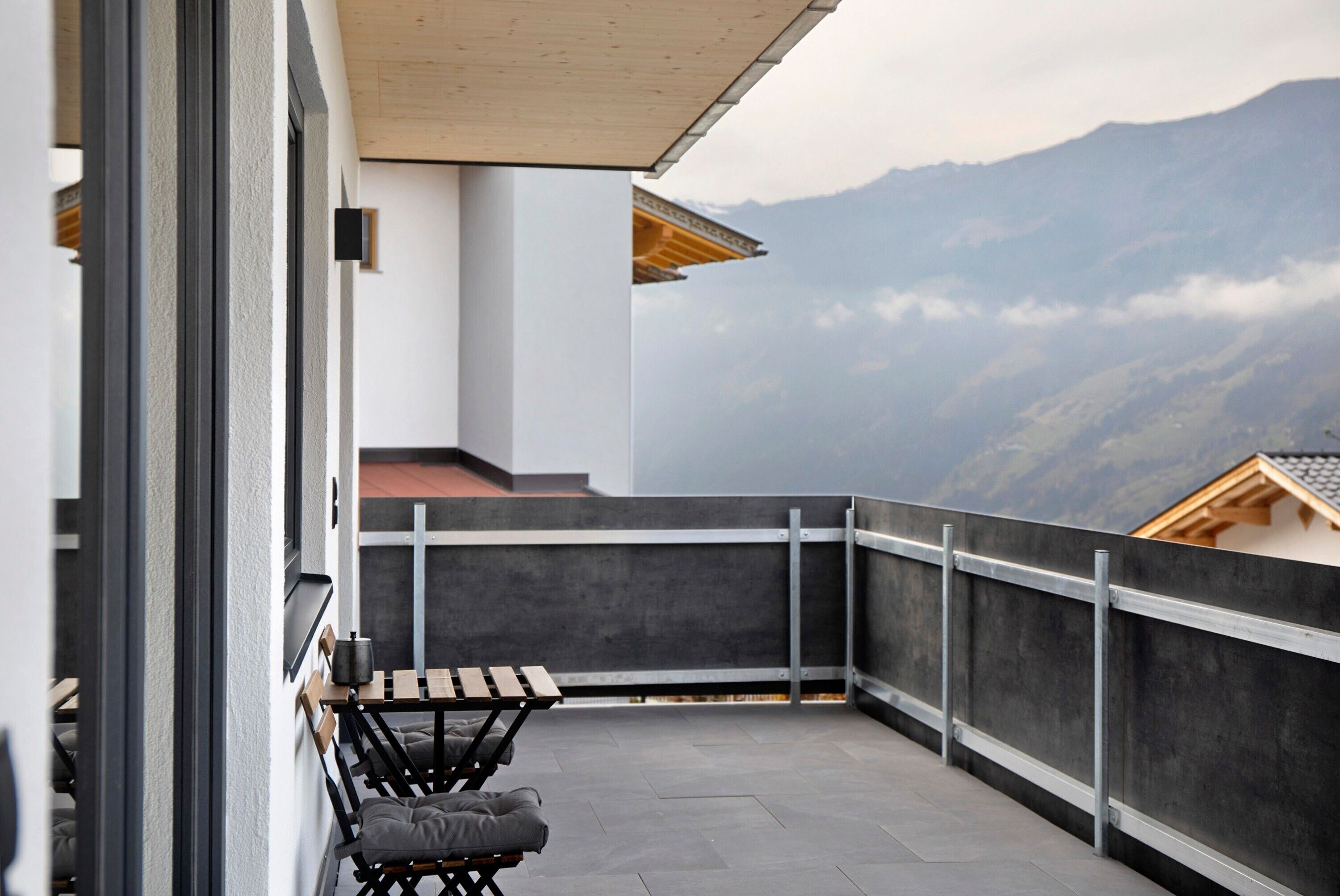 A balcony with a table and chairs, a view of mountains, and a modern building facade.